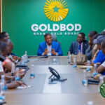 Executive boardroom briefing with a man in a blue suit speaking at the head of a long table, surrounded by diverse attendees; a green wall with the GOLD BOD/Ghana Gold Board logo behind him.