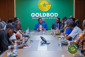 Executive boardroom briefing with a man in a blue suit speaking at the head of a long table, surrounded by diverse attendees; a green wall with the GOLD BOD/Ghana Gold Board logo behind him.