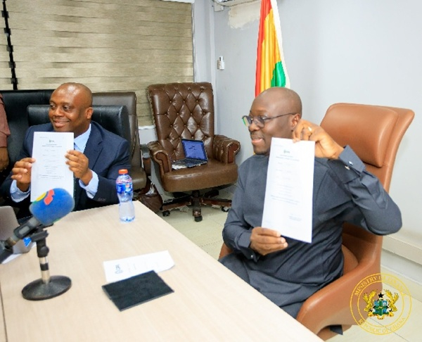 Two men in suits sit at a conference table, each holding up a document as a microphone and water bottle sit nearby, suggesting a press briefing or signing event.