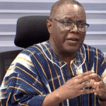 Man in blue and white striped traditional attire, wearing glasses, speaking and gesturing with his hands in an office setting.