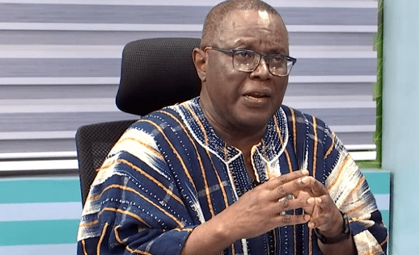 Man in blue and white striped traditional attire, wearing glasses, speaking and gesturing with his hands in an office setting.