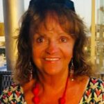 Smiling woman with shoulder-length brown hair wearing a colorful patterned top and a red bead necklace at an outdoor cafe.
