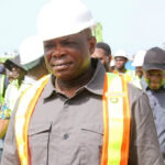 Construction site supervisor in a white hard hat and orange safety vest leads a group of workers on a construction site.