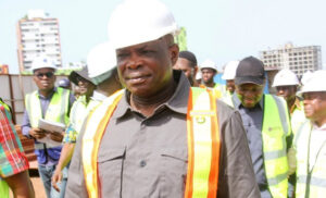 Construction site supervisor in a white hard hat and orange safety vest leads a group of workers on a construction site.