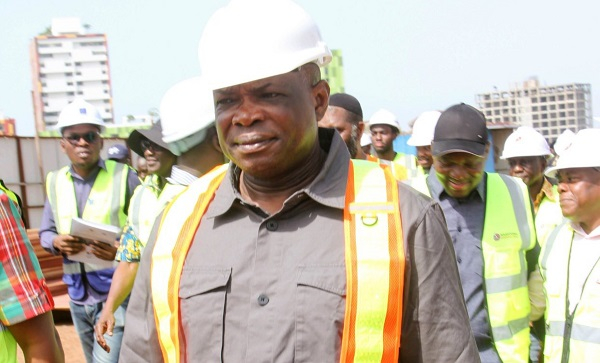 Construction site supervisor in a white hard hat and orange safety vest leads a group of workers on a construction site.