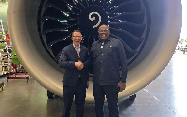 Two men shake hands in front of a gigantic jet engine inside a hangar, one in a suit and the other in dark work attire, both smiling at the camera.