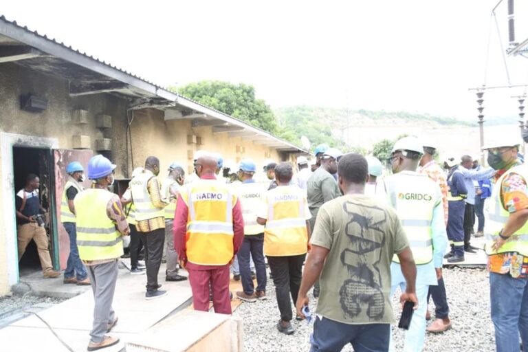 Group of people in safety vests and helmets standing together at a construction site beside a single-story building with gravel ground.