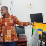 Man in a colorful patchwork shirt giving a presentation, pointing at a monitor showing a yellow grid/map on a desk in an office setting
