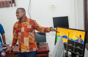 Man in a colorful patchwork shirt giving a presentation, pointing at a monitor showing a yellow grid/map on a desk in an office setting