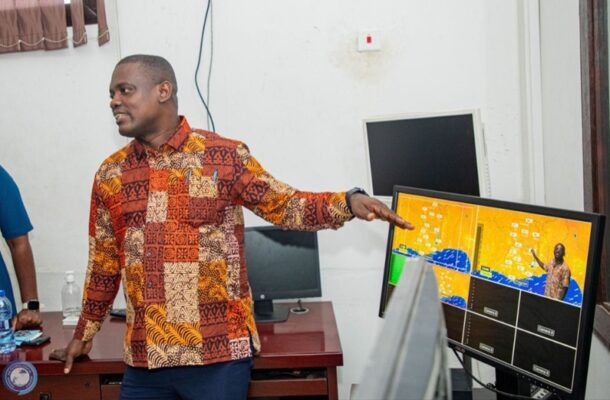 Man in a colorful patchwork shirt giving a presentation, pointing at a monitor showing a yellow grid/map on a desk in an office setting