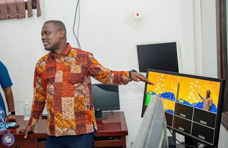 Man in a colorful patchwork shirt giving a presentation, pointing at a monitor showing a yellow grid/map on a desk in an office setting
