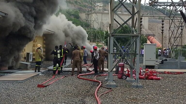 Firefighters in protective gear fight a fire at an industrial site, thick black smoke pouring from a building as red hoses snake across gravel and workers watch.