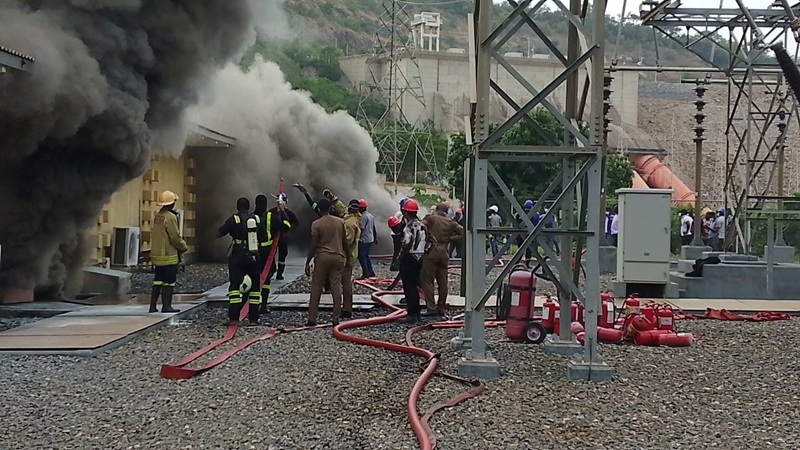 Firefighters in protective gear fight a fire at an industrial site, thick black smoke pouring from a building as red hoses snake across gravel and workers watch.
