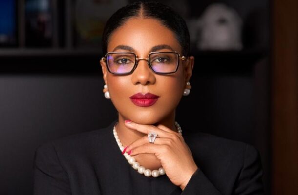 Professional woman with dark hair and glasses, wearing a pearl necklace and earrings, resting her chin on her hand during a formal portrait.