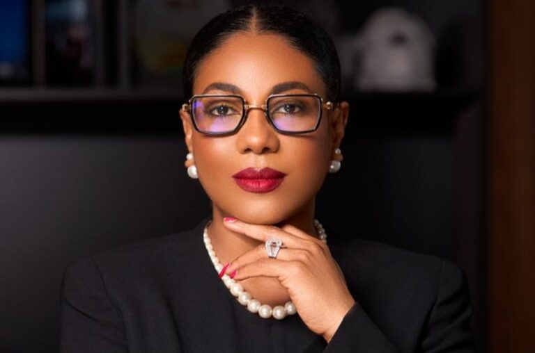 Professional woman with dark hair and glasses, wearing a pearl necklace and earrings, resting her chin on her hand during a formal portrait.