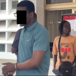 Outdoor scene with two people outside a building: a man in a teal polo in the foreground with hands clasped, and a woman in an orange shirt with a crossbody bag near a red notice board in the background.