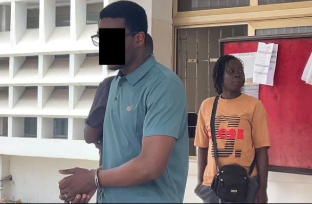 Outdoor scene with two people outside a building: a man in a teal polo in the foreground with hands clasped, and a woman in an orange shirt with a crossbody bag near a red notice board in the background.