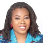 Portrait of a smiling woman with dreadlocks and red earrings, wearing a blue patterned blazer.