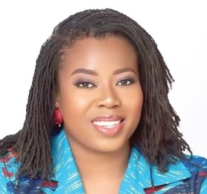 Portrait of a smiling woman with dreadlocks and red earrings, wearing a blue patterned blazer.