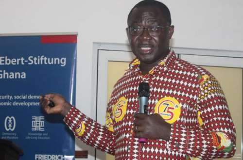 Speaker in a red-and-white patterned shirt, holding a microphone and gesturing, with a yellow 75 badge, beside a blue Ebert-Stiftung Ghana banner.