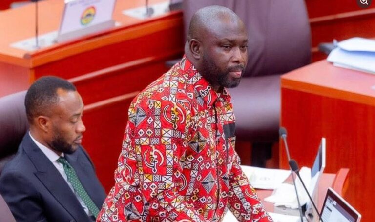 Man in a red patterned shirt speaks at a podium in a formal meeting room, with a colleague seated beside him.