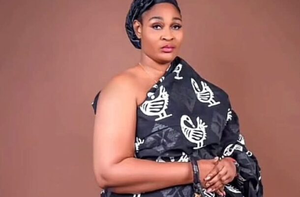 Woman in a black off-shoulder dress with white bird print, wearing a headwrap, posing with hands folded in front against a brown backdrop.