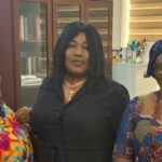 Three women posing together indoors; middle woman in black with a cross necklace flanked by women in colorful head wraps and patterned outfits.