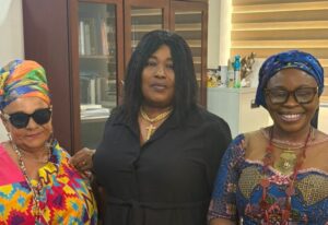 Three women posing together indoors; middle woman in black with a cross necklace flanked by women in colorful head wraps and patterned outfits.