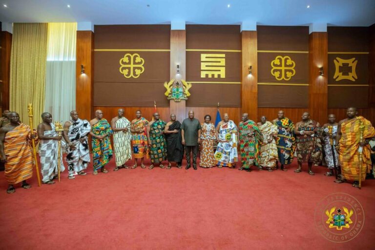 Group of Ghanaian officials in colorful traditional robes standing in a formal hall with the presidential crest in the background.