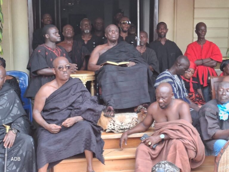 Group of men in dark robes gathers on building steps; central elder holds an open book while others sit or stand around.
