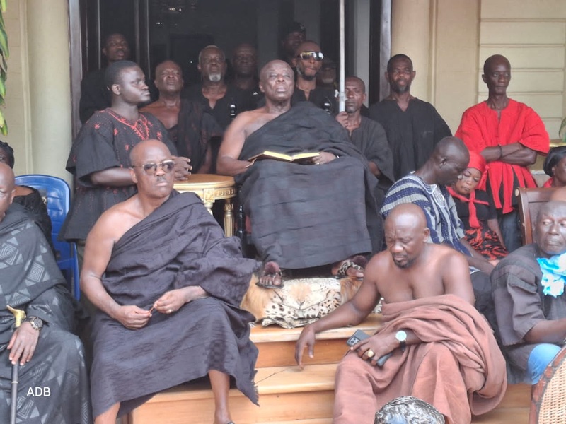 Group of men in dark robes gathers on building steps; central elder holds an open book while others sit or stand around.