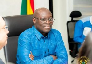 Man with glasses in a blue shirt sits at a conference table during a meeting, with a flag and office chairs in the background.