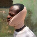Side profile of a man wearing a beige cloth hood wrapped around his head and chin, beside a weathered green wall.