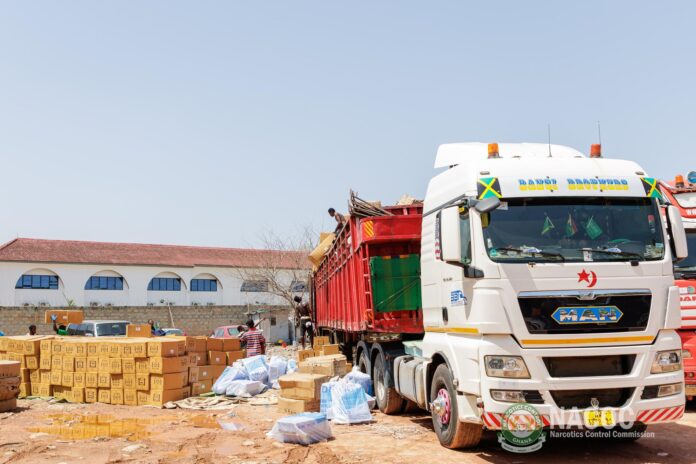 White cargo truck at a dusty loading yard with boxes and bags stacked nearby; workers handle cargo.