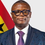 Portrait of a man in a blue patterned suit with a white shirt and purple tie, wearing blue glasses, standing in front of the Ghana flag.