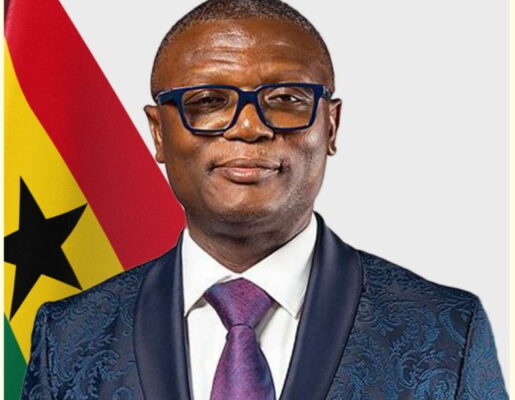IMG_0119 | Ghanaguardian.com Portrait of a man in a blue patterned suit with a white shirt and purple tie, wearing blue glasses, standing in front of the Ghana flag.