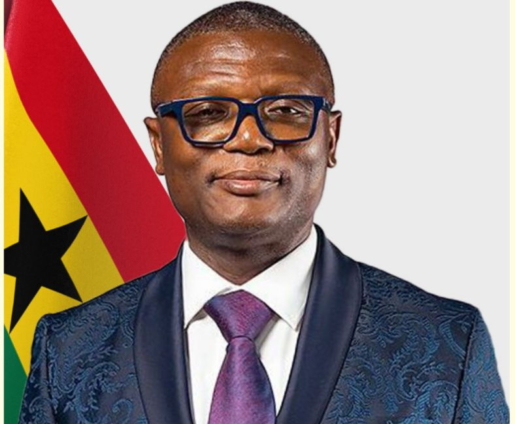 Portrait of a man in a blue patterned suit with a white shirt and purple tie, wearing blue glasses, standing in front of the Ghana flag.