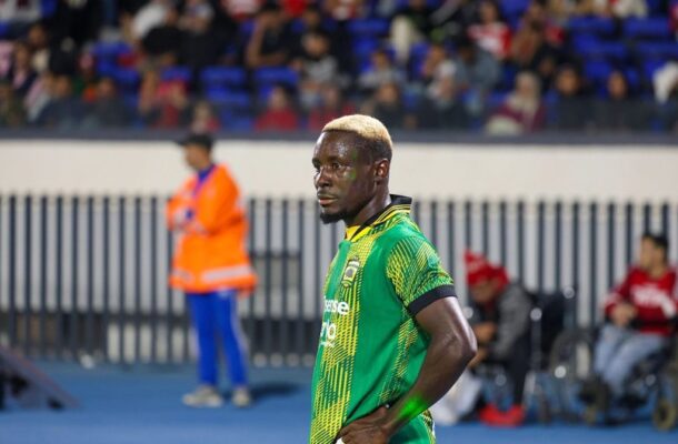 Soccer player in a green jersey standing on the blue field with spectators in the stands behind him.