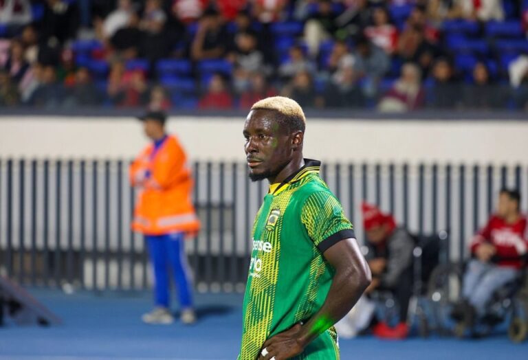 Soccer player in a green jersey standing on the blue field with spectators in the stands behind him.