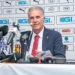 Older man in a dark suit and red tie speaks at a press conference, microphones in front of him and sponsor logos behind.