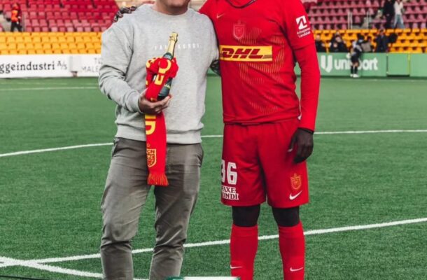 Football player in red kit poses with a man in a grey sweatshirt on a stadium pitch, holding a champagne bottle and scarf. A green Faxe Kondi box sits on the ground nearby.