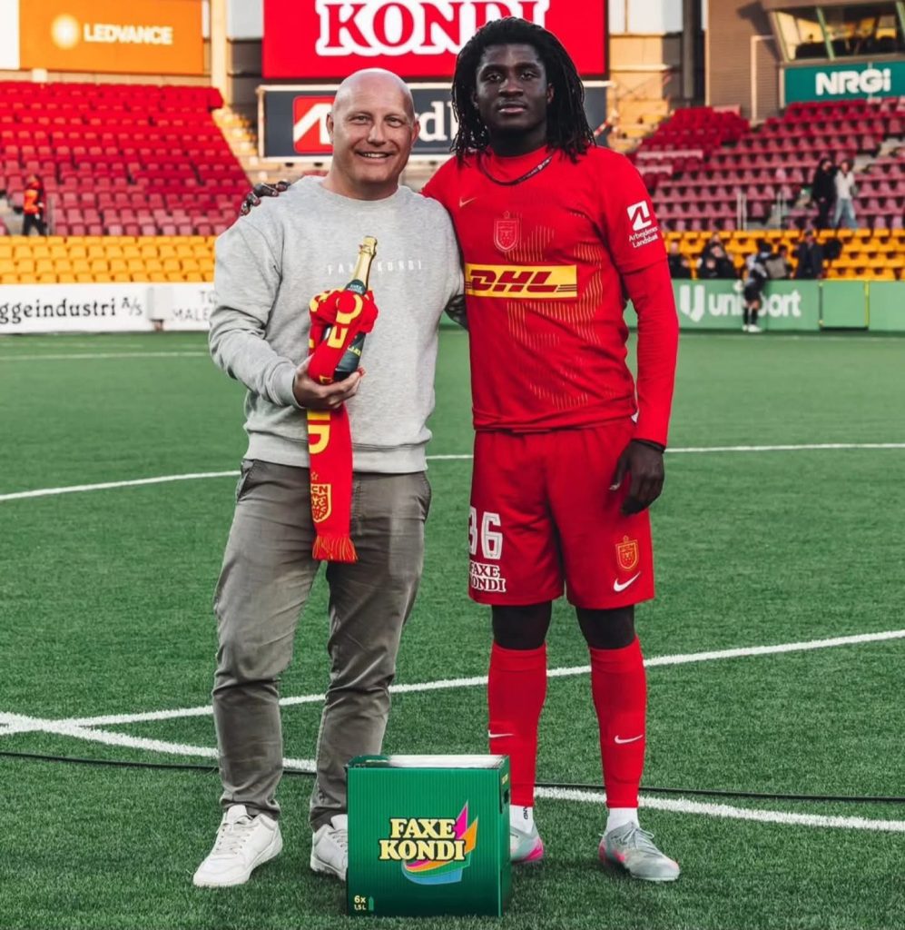Football player in red kit poses with a man in a grey sweatshirt on a stadium pitch, holding a champagne bottle and scarf. A green Faxe Kondi box sits on the ground nearby.