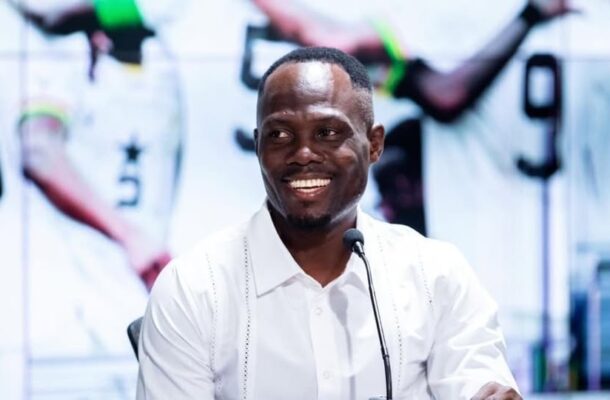 Smiling man in a white shirt speaks into a microphone at a panel or press event, with blurred figures in the background behind him.
