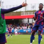 Soccer players celebrate on a green field; a player in a blue-and-red kit runs toward a teammate with arms raised as others cheer in the background.