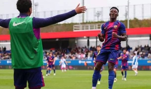 Soccer players celebrate on a green field; a player in a blue-and-red kit runs toward a teammate with arms raised as others cheer in the background.