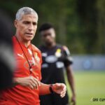 Coach with gray hair in a red training shirt giving instructions on a football practice field, players in black jerseys in the background