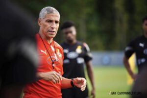 Coach with gray hair in a red training shirt giving instructions on a football practice field, players in black jerseys in the background