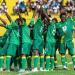 Soccer team in green uniforms celebrates in a circle on the field with raised arms, yellow stands behind them.