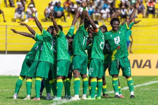 Soccer team in green uniforms celebrates in a circle on the field with raised arms, yellow stands behind them.