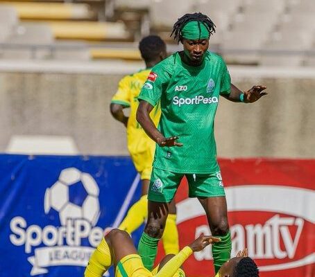 Soccer player in a green uniform stands over a fallen opponent in yellow on a grassy field.